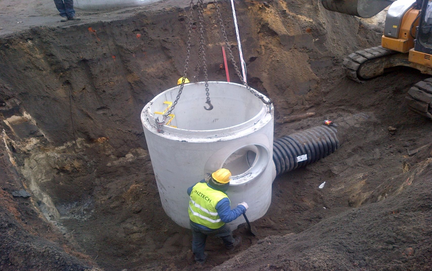 Montaż betonowego kręgu studni kanalizacyjnej z podłączeniem rury karbowanej w wykopie ziemnym, widoczny pracownik z łopatą i koparka w tle.