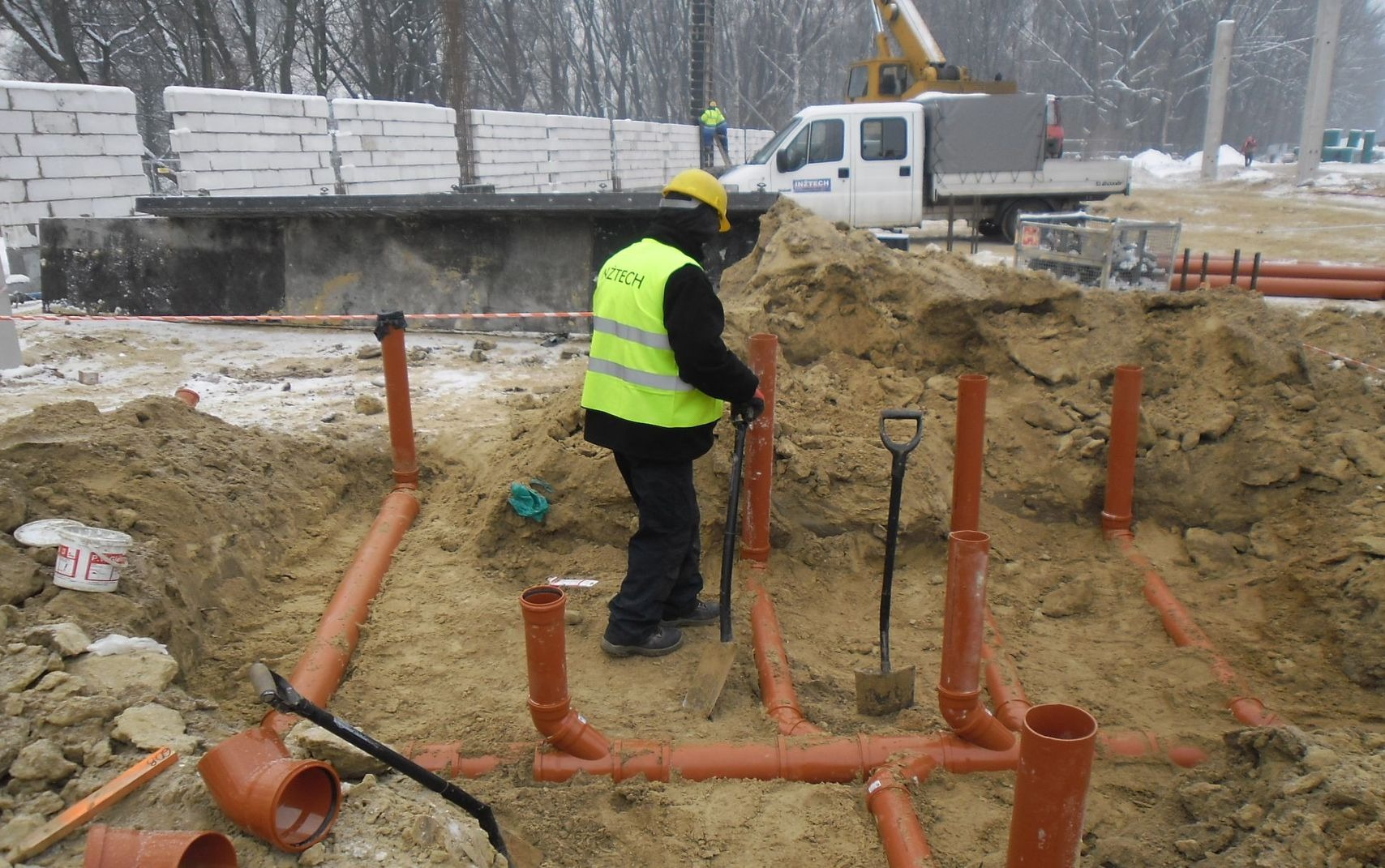 Pracownik w żółtej kamizelce i kasku montuje pomarańczowe rury kanalizacyjne w wykopie na placu budowy, widoczny sprzęt budowlany i śnieg.