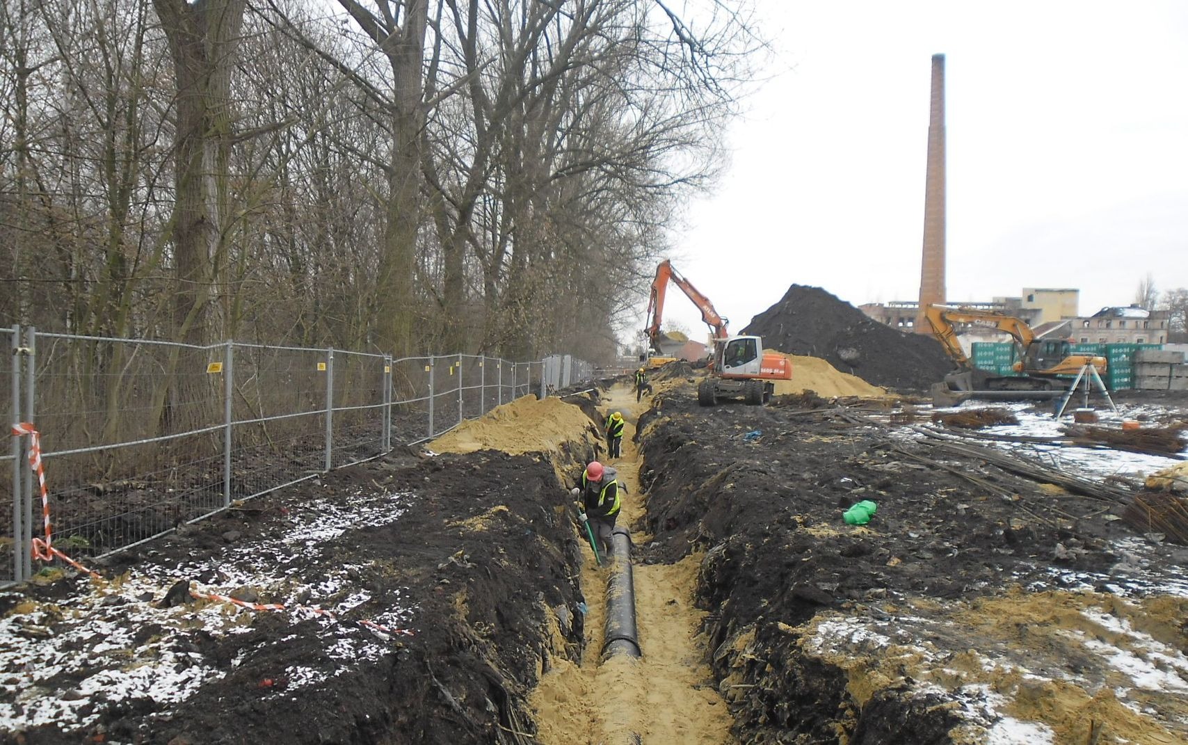 Montaż rury w wykopie, pracownicy w odblaskowych kamizelkach, w tle koparki i ceglany komin, teren budowy zabezpieczony siatką.