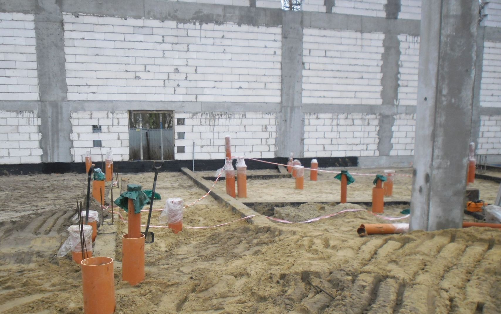 Wykop z wystającymi pionowymi rurami kanalizacyjnymi zabezpieczonymi folią i tkaniną, na tle ściany z białych bloczków betonowych i betonowych słupów nośnych.