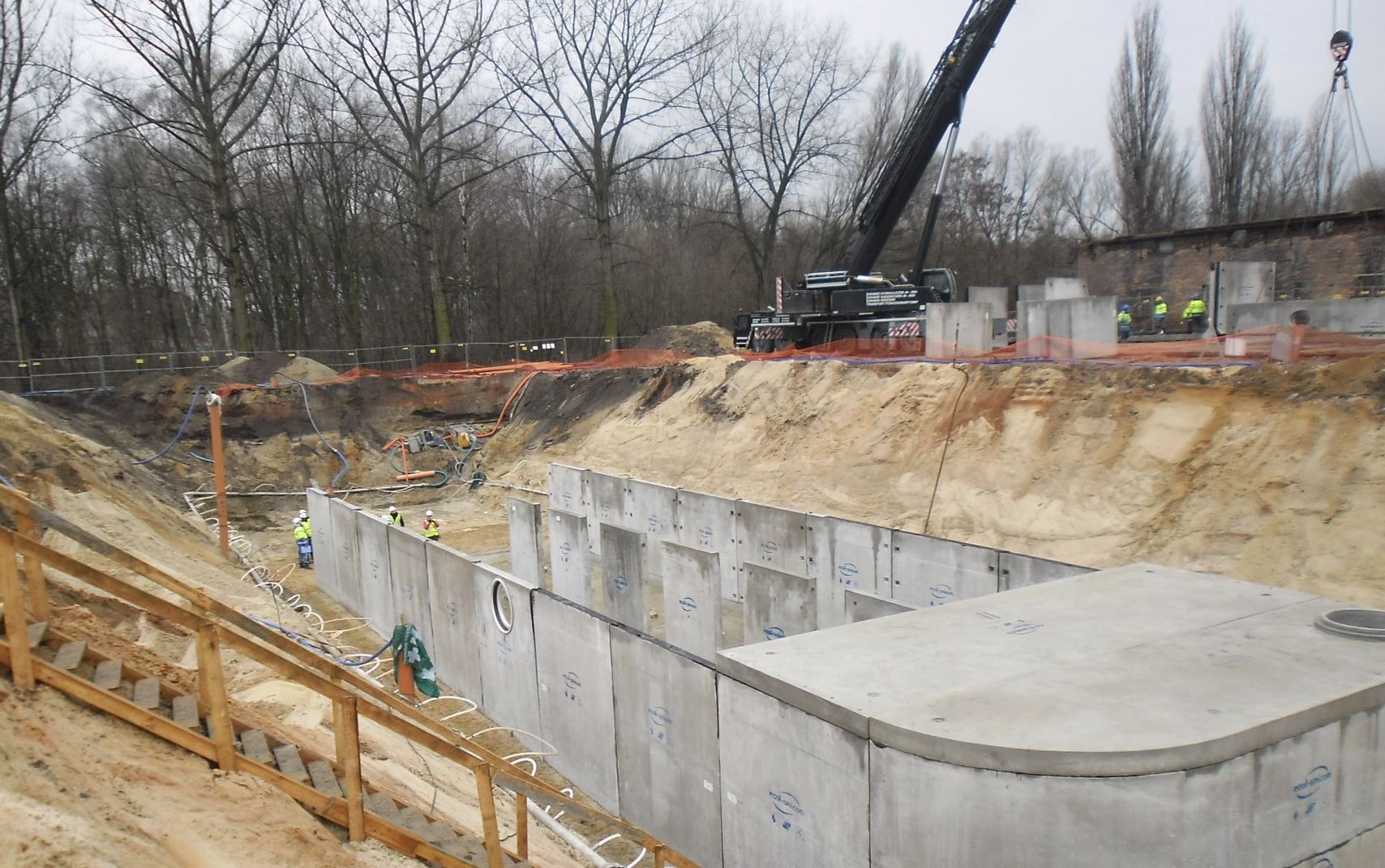 Montaż prefabrykowanego betonowego zbiornika podziemnego na placu budowy, widoczny dźwig i pracownicy w odblaskowych kamizelkach.