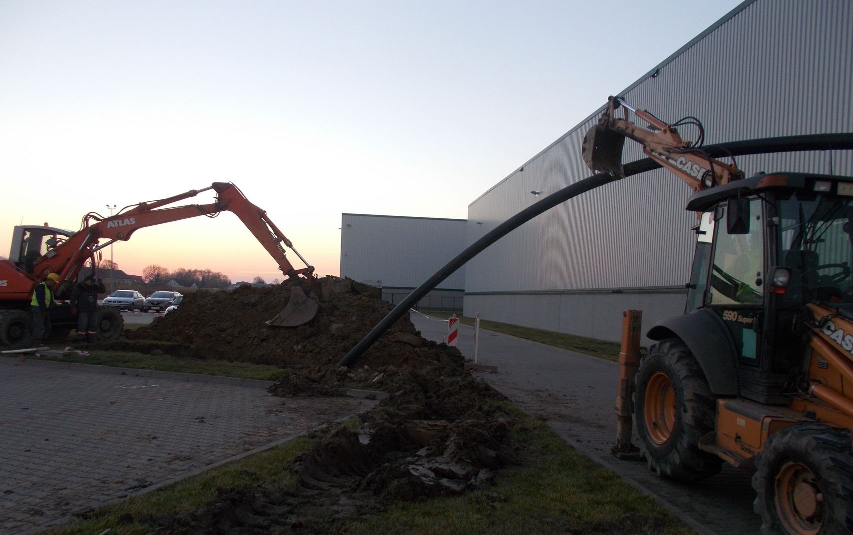 Dwa pomarańczowe traktory z łyżkami układają czarną, grubą rurę w wykopie ziemnym na tle budynku przemysłowego o blaszanej elewacji, widoczne znaki ostrzegawcze i zaparkowane samochody w oddali...