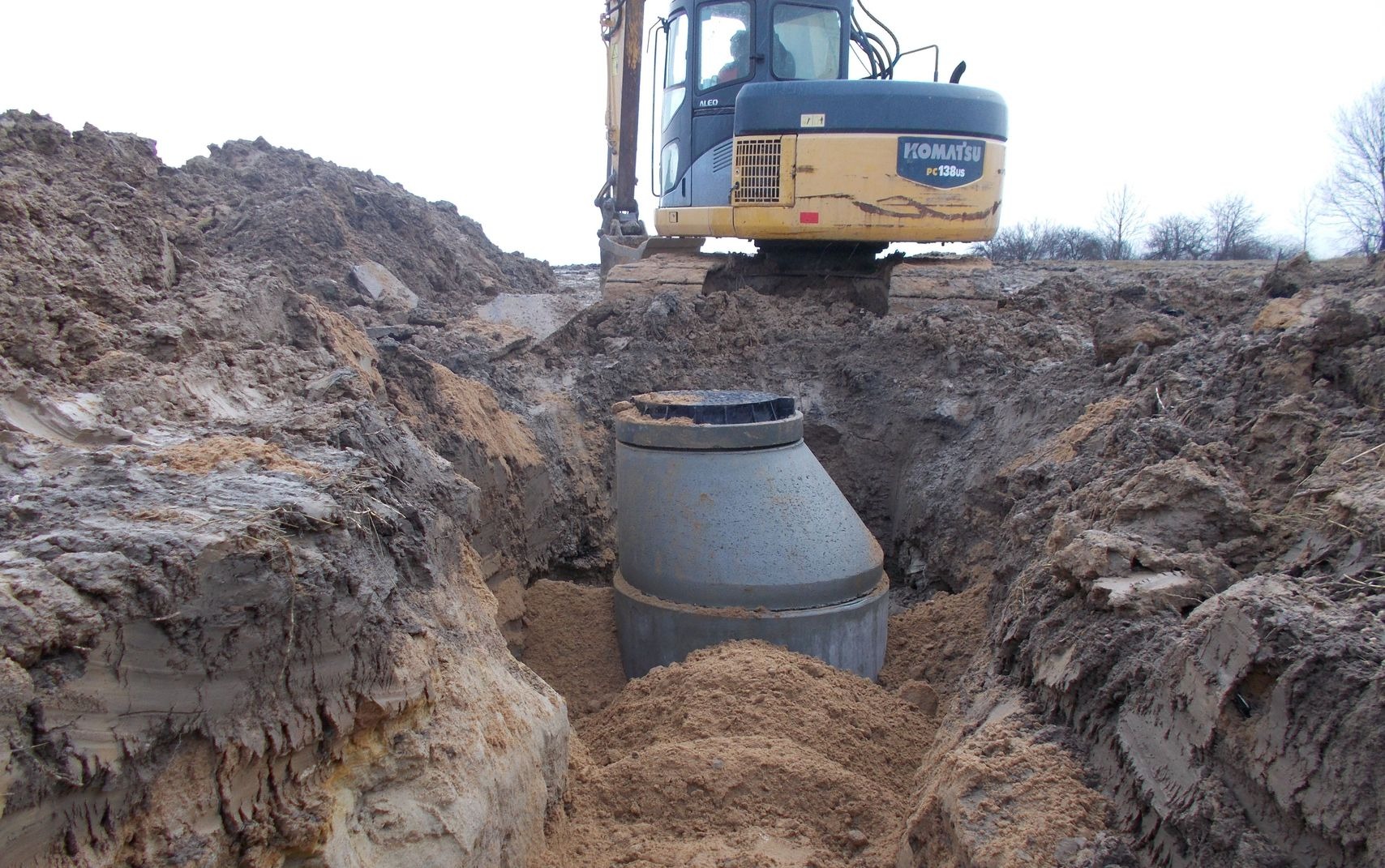 Betonowy zbiornik z czarną pokrywą umieszczony w wykopie, z koparką Komatsu PC138US w tle, podczas prac ziemnych.