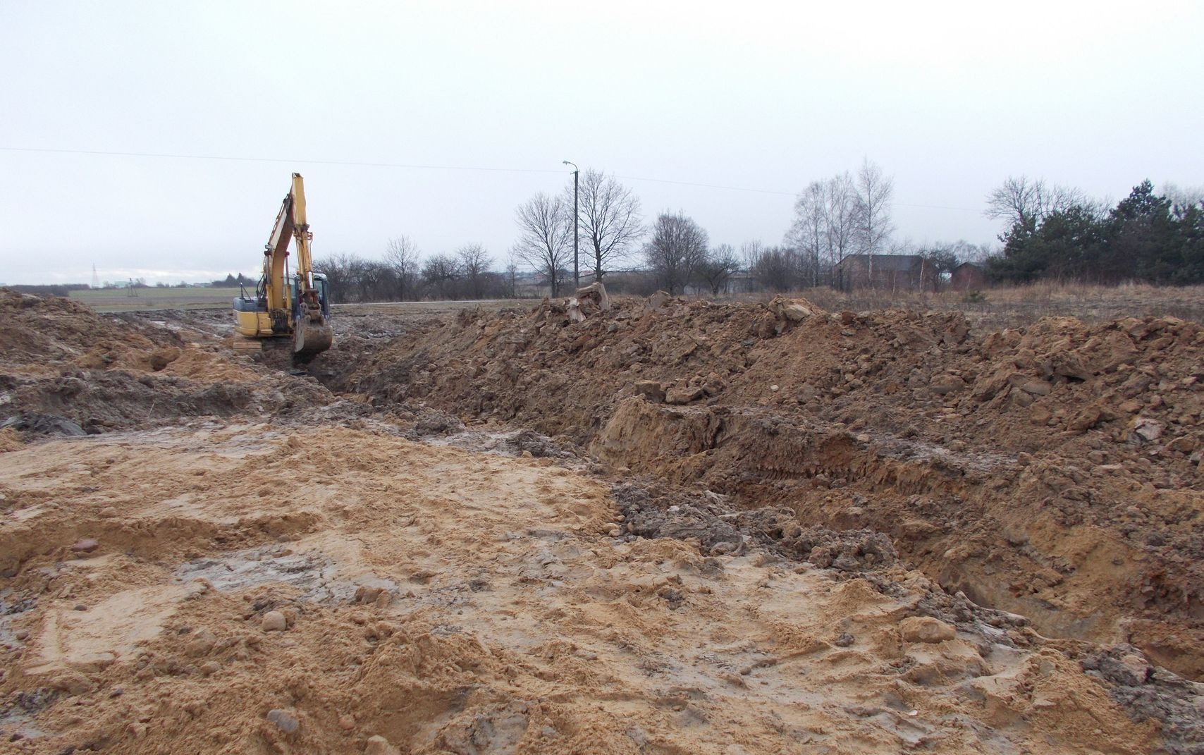 Żółta koparka głębi w wykopie ziemnym na tle pochmurnego nieba i drzew bez liści.