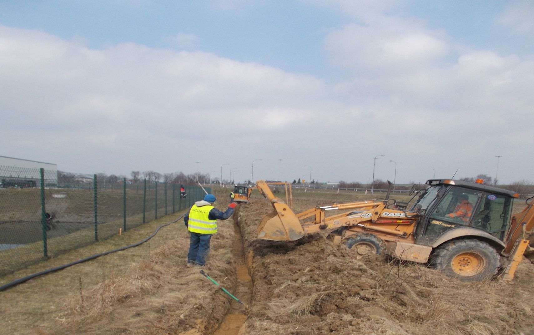 Żółta koparko-ładowarka Case 580 Super R wykopuje rów na polu, pracownik w żółtej kamizelce nadzoruje prace, w tle widoczna autostrada i zabudowania.