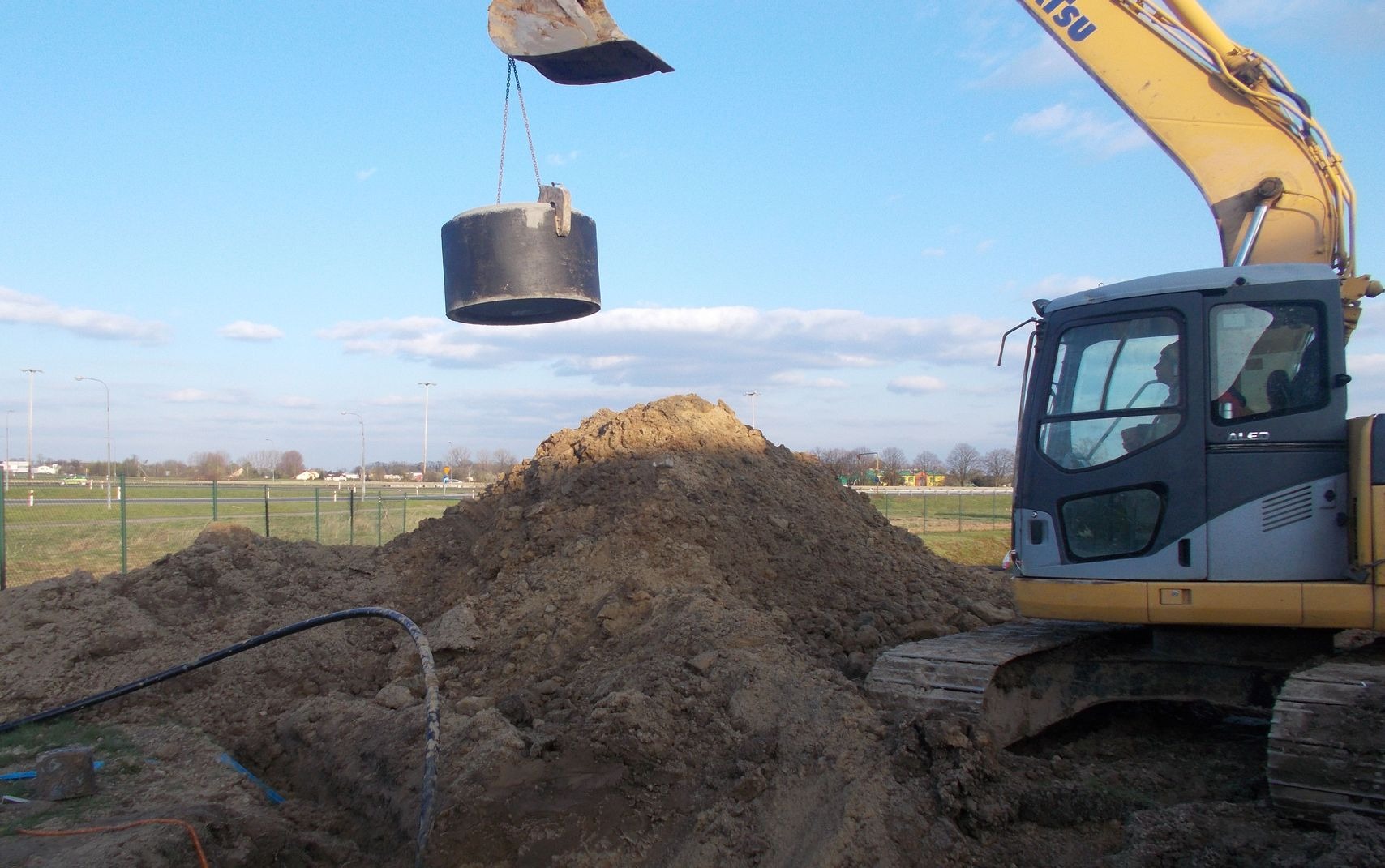 Żółto-szara koparka Komatsu z operatorem w kabinie, podnosi cylindryczny element nad stertą ziemi, widoczne rury w wykopie, w tle ogrodzenie, latarnie i budynki.