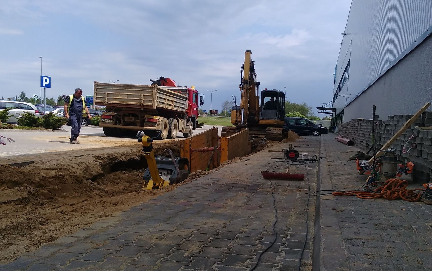 Wykop zabezpieczony szalunkiem stalowym, koparka, ubijarka i ciężarówka na tle budynku przemysłowego, robotnik z poziomicą.