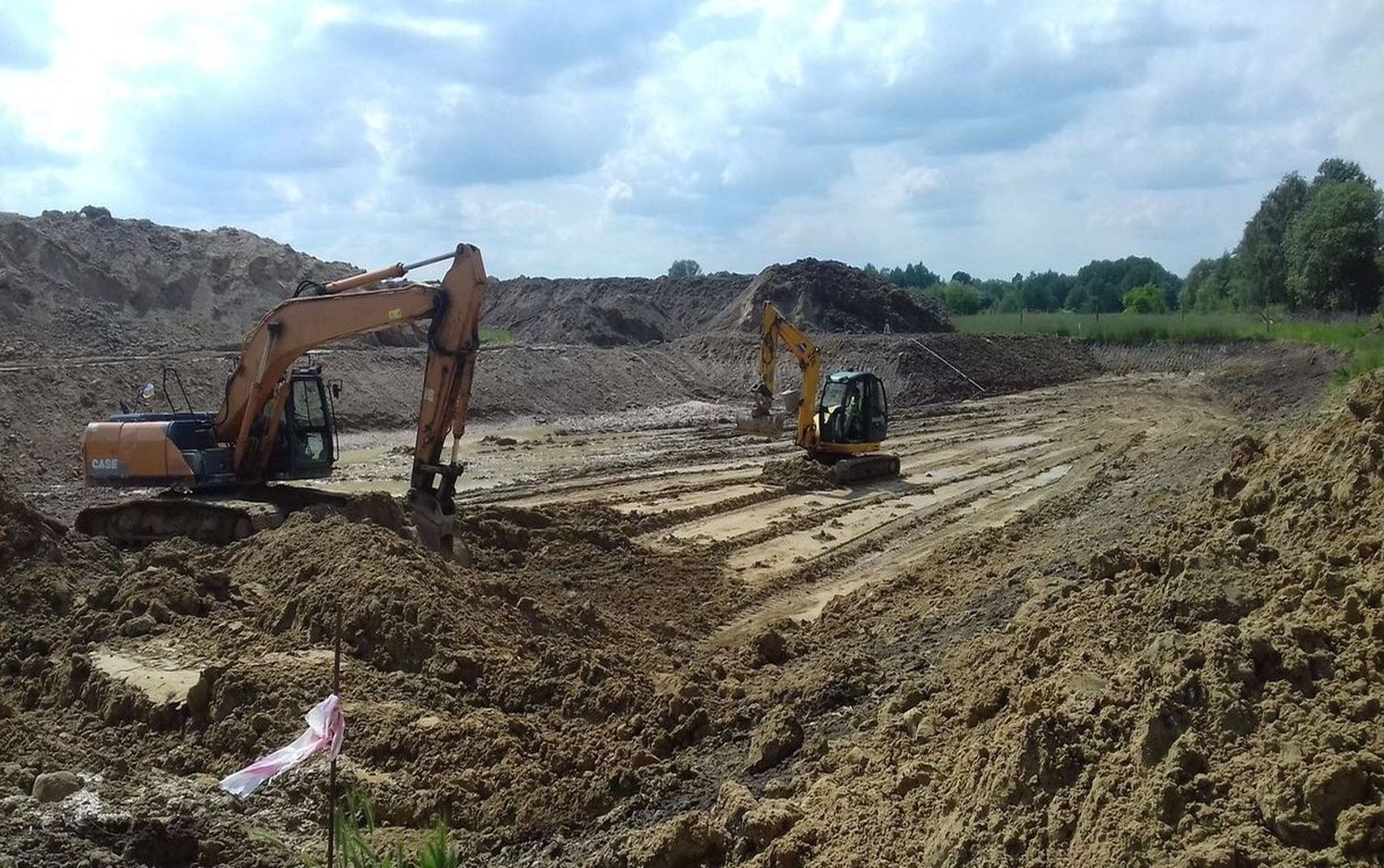 Dwie koparki Case pracują przy wykopie zbiornika wodnego na tle pochmurnego nieba, z widocznymi śladami opon na ziemi i plastikową taśmą ostrzegawczą w pobliżu jednej z maszyn.