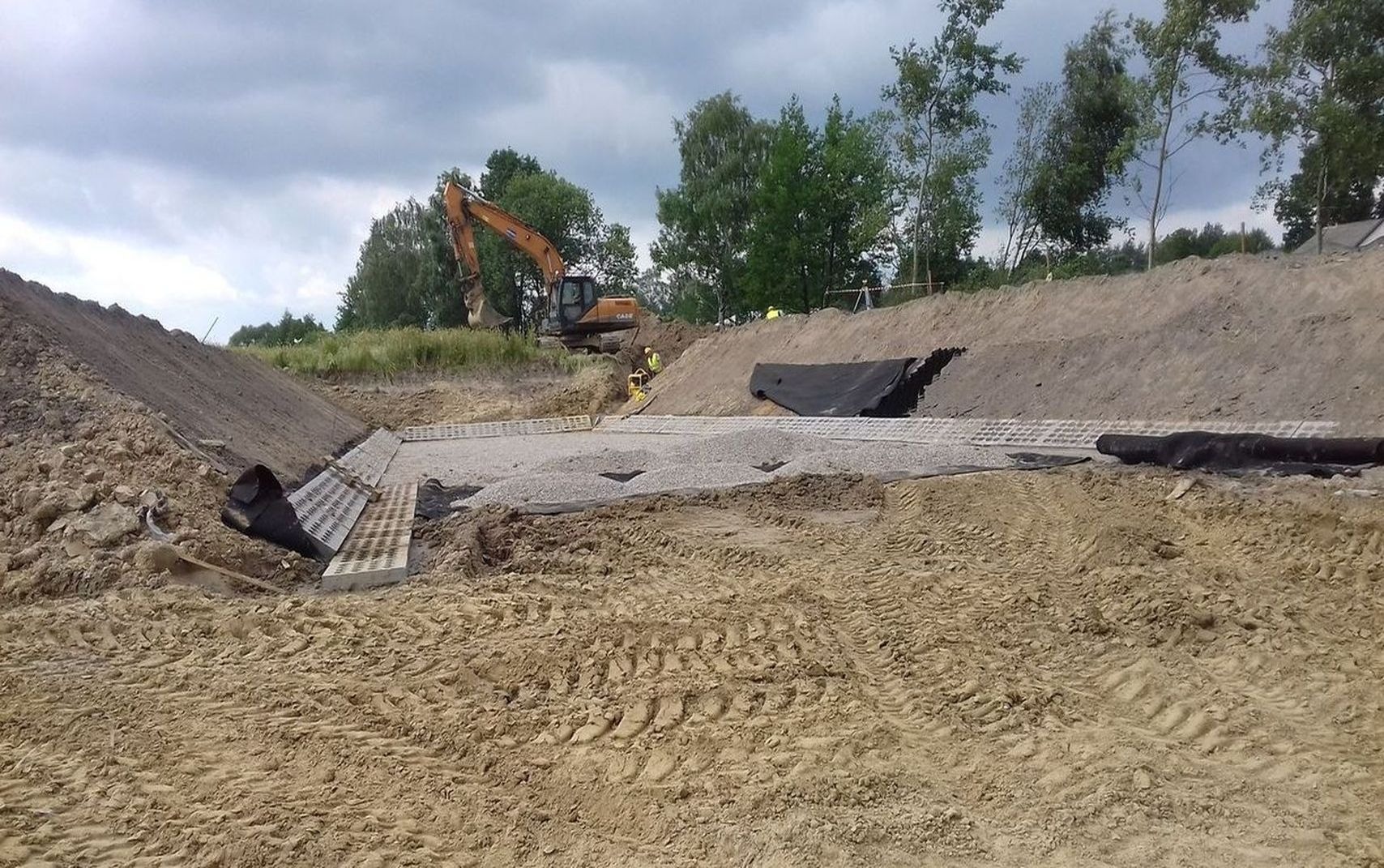 Budowa przepustu drogowego: Wykop z ułożonymi prefabrykatami betonowymi, warstwą żwiru i czarną geowłókniną, w tle koparka i pracownicy w żółtych kamizelkach.