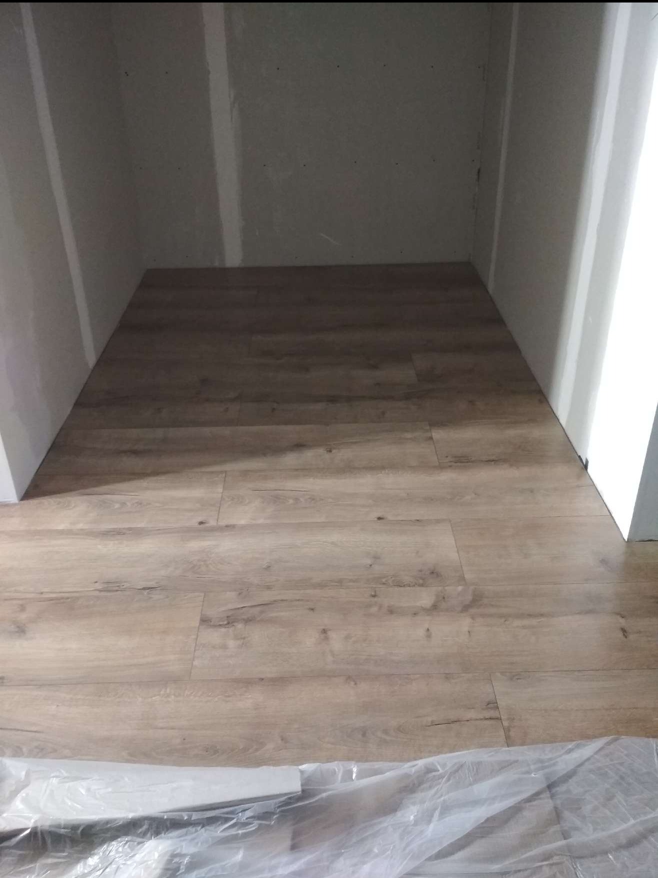 Ułożone panele podłogowe w małym pomieszczeniu z surowymi ścianami. Widoczne panele podłogowe i fragment folii ochronnej na podłodze.