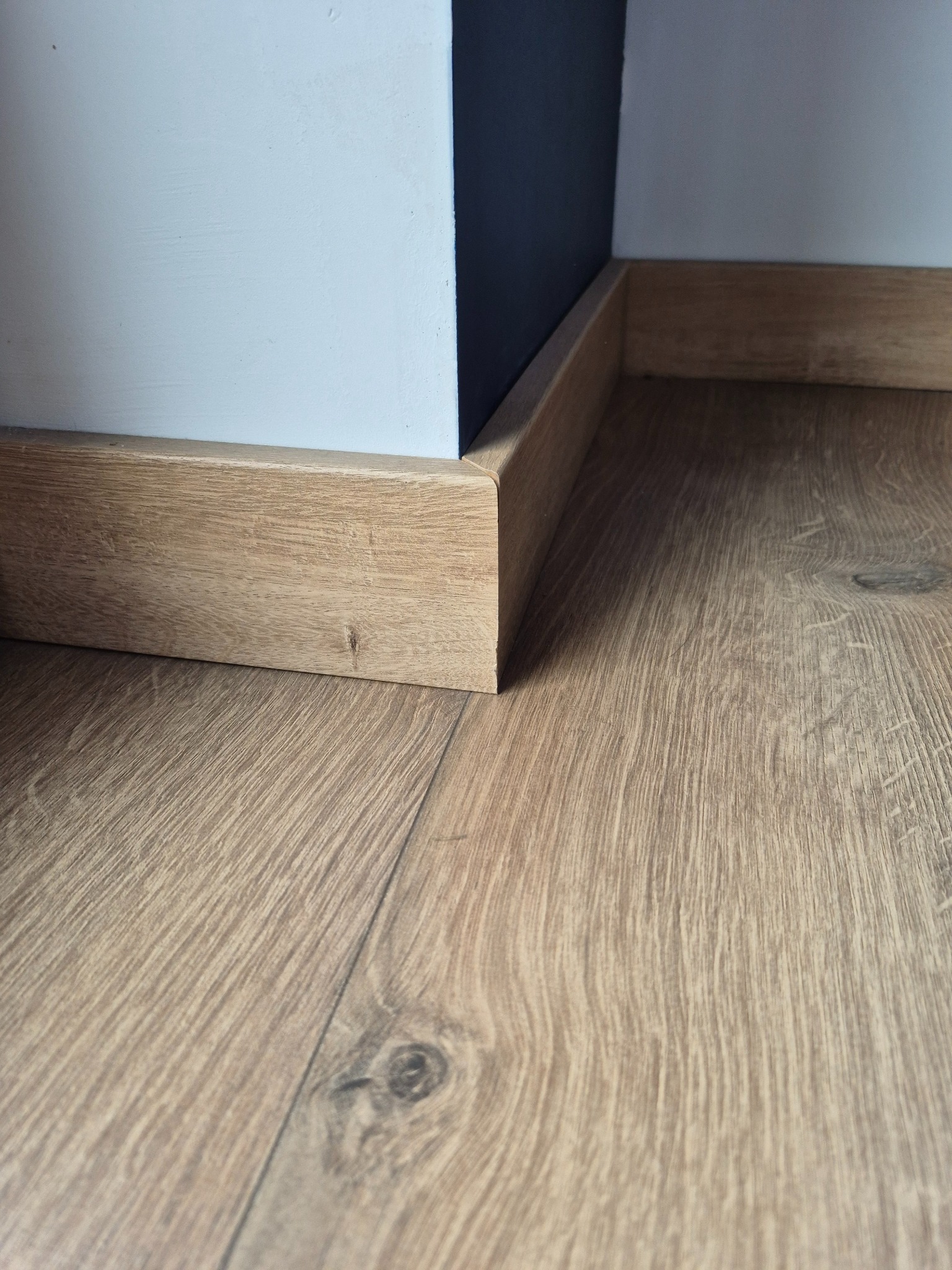 Jasne panele podłogowe z drewnopodobnym wzorem, przy ścianie z zamontowaną listwą przypodłogową w kolorze podłogi, widok z bliska na łączenie paneli i listwy.