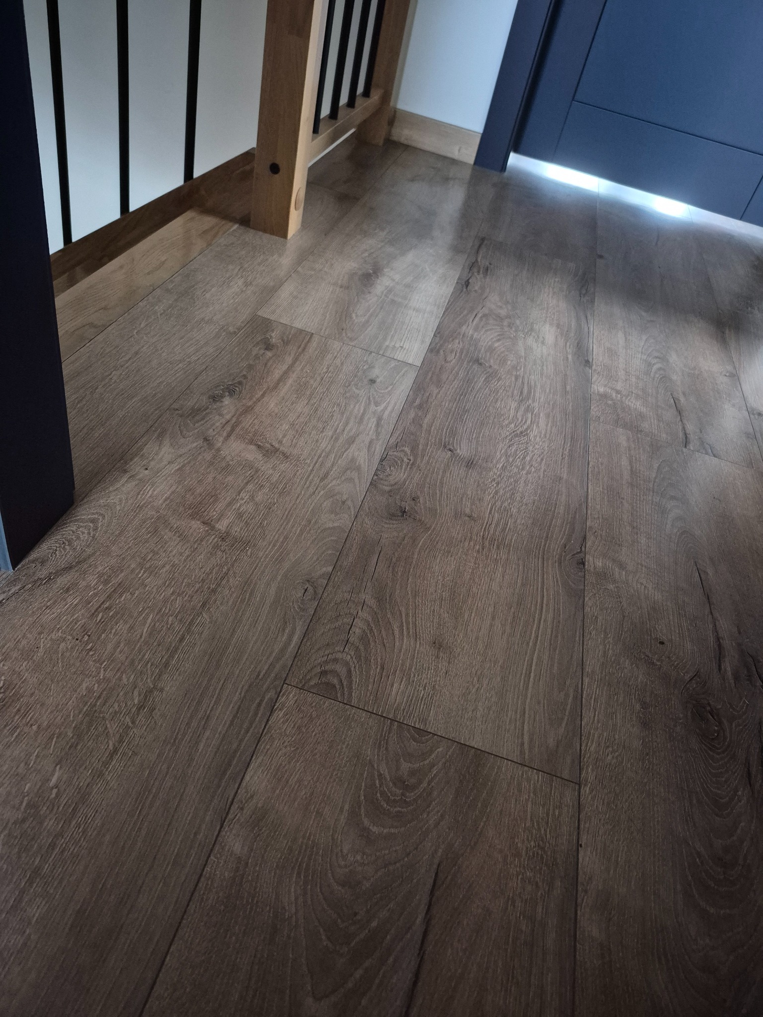 Ułożone panele podłogowe w odcieniach brązu, widoczne przy schodach i drzwiach. Naturalne oświetlenie podkreśla strukturę drewna.