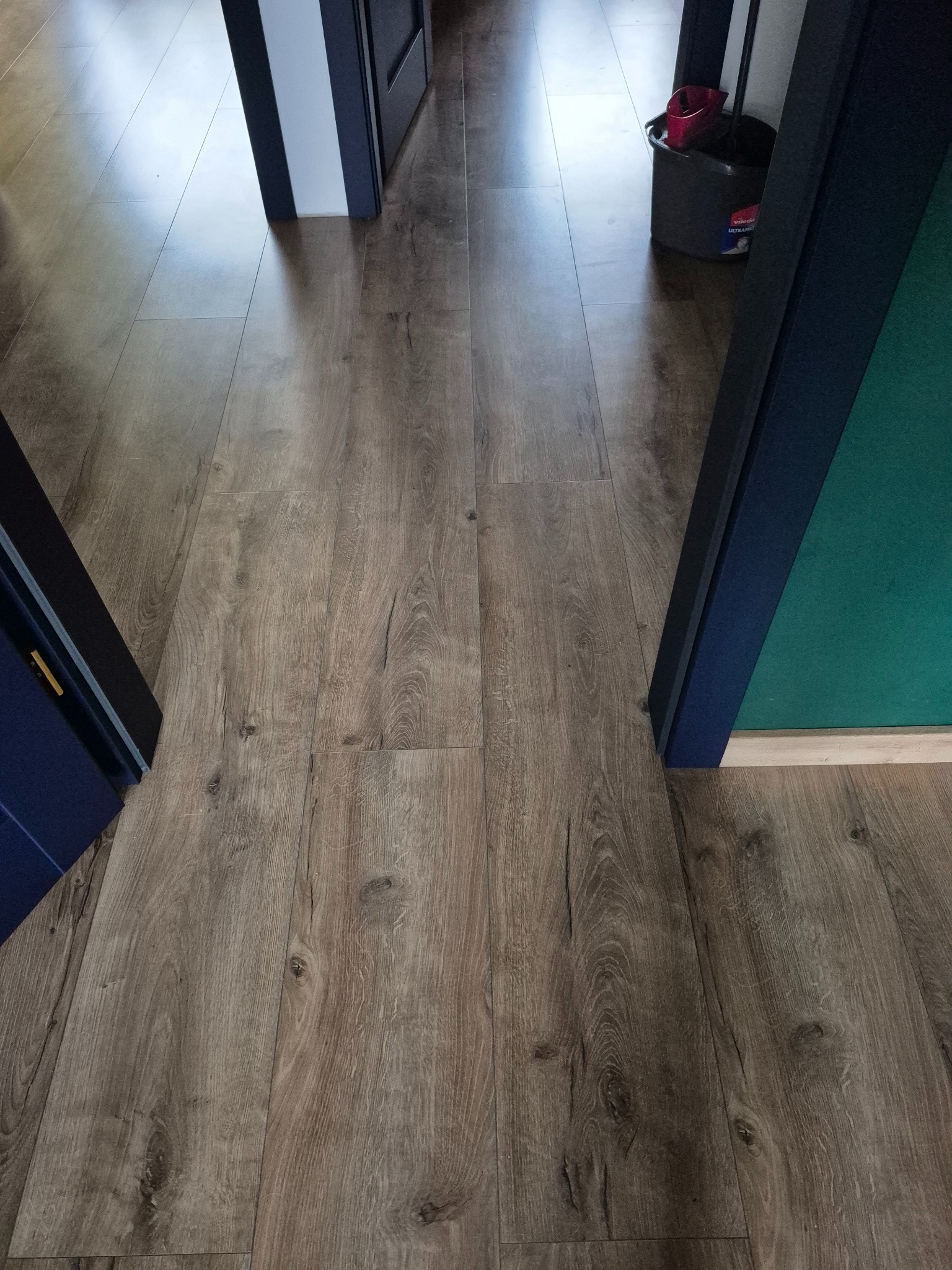 Ułożone panele podłogowe w odcieniach brązu, widoczne fragmenty ościeżnic w kolorze granatowym i białym. W tle wiadro z mopem.