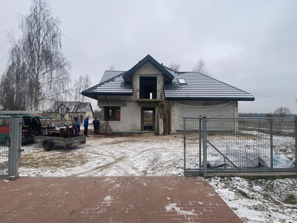 Dom w trakcie budowy z częściowo ukończonym dachem pokrytym szarą dachówką, widoczne elementy konstrukcyjne i materiały budowlane, na zewnątrz ekipa budowlana i przyczepka z elementami drewnianymi,...