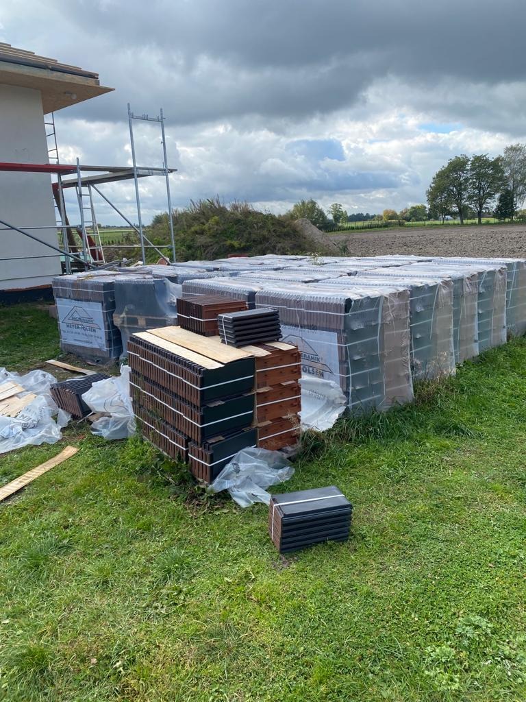 Palety z dachówkami ceramicznymi w folii ochronnej, ułożone na trawie obok budynku w trakcie budowy, pochmurne niebo w tle.