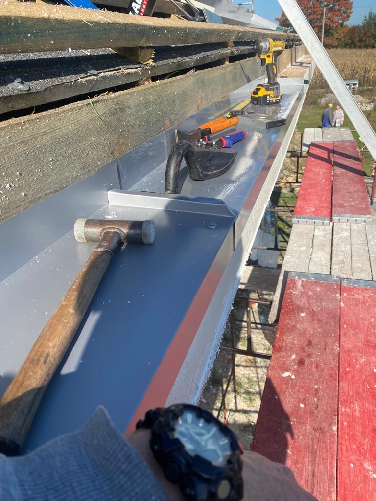 Montaż nowej, metalowej rynny dachowej z widocznymi narzędziami: młotek, wkrętarka akumulatorowa, szczypce i poziomica, na tle rusztowania i pola.