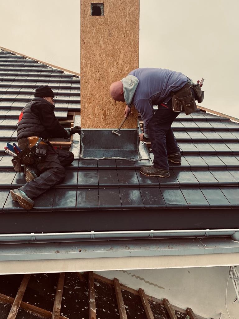 Dwóch dekarzy pracuje nad obróbką blacharską komina na dachu pokrytym ciemną dachówką. Jeden dekarz przytrzymuje element, drugi wbija go młotkiem. Widoczna więźba dachowa od spodu.