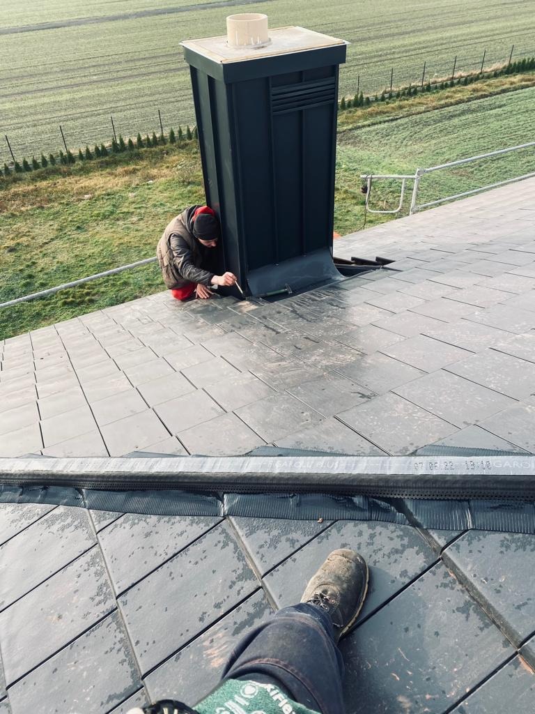 Dekarz uszczelnia obróbkę blacharską komina na dachu pokrytym szarą dachówką, widok z perspektywy osoby stojącej na dachu.