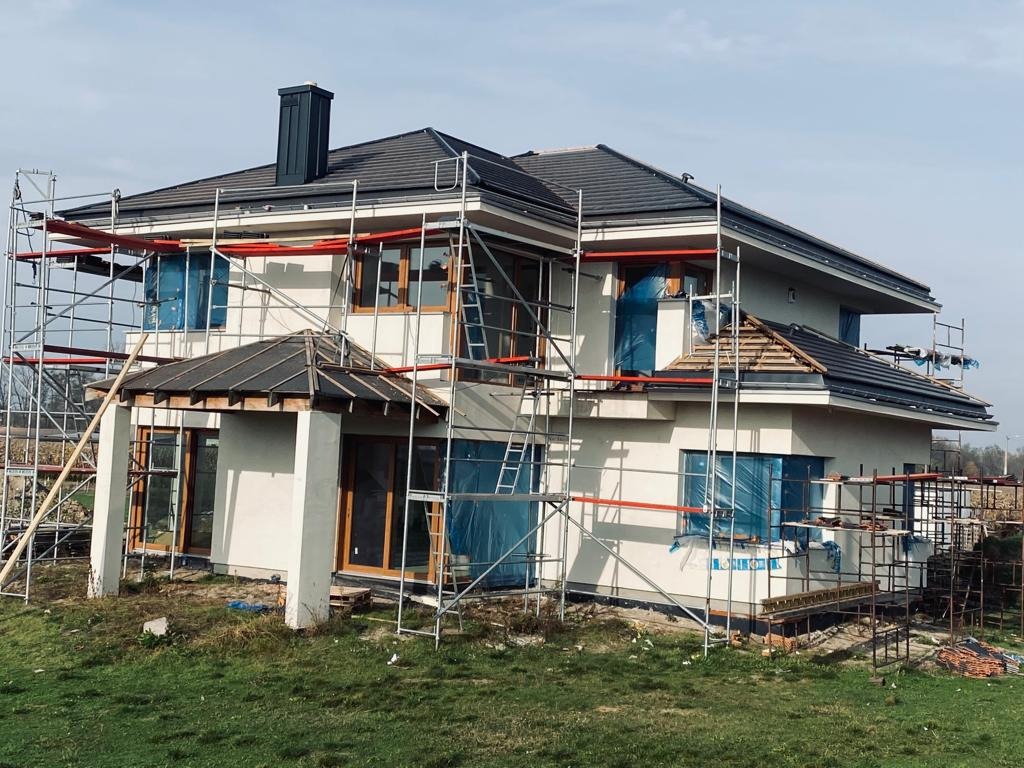 Dom w trakcie prac dekarskich, widoczne rusztowania, częściowo położone ciemne dachówki i zabezpieczone okna folią.