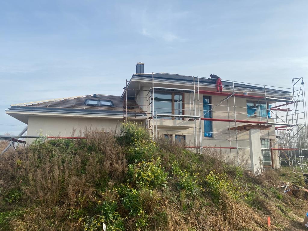 Dwukondygnacyjny dom w trakcie prac dekarskich; widoczne rusztowania, częściowo ułożona dachówka i dekarz na dachu.