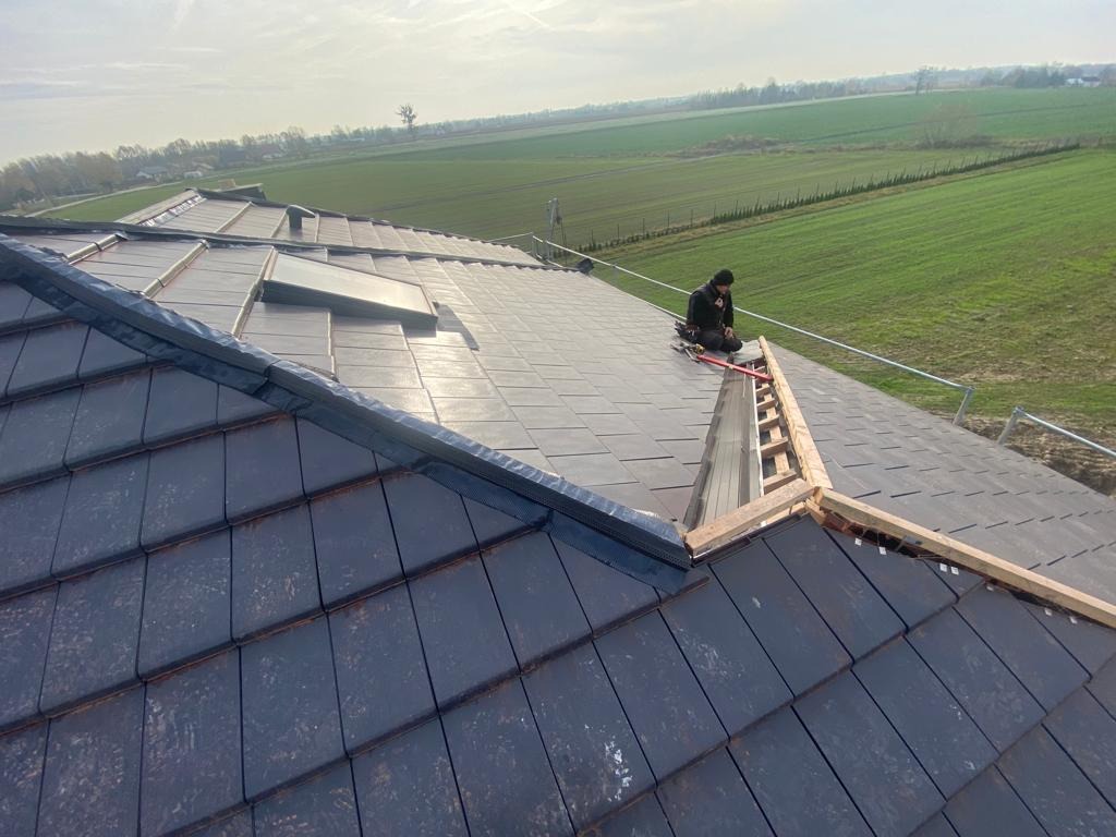 Dekarz na dachu z ciemnej dachówki ceramicznej, montujący obróbki blacharskie przy oknie dachowym, widok na pola i drzewa w oddali.