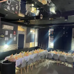 Uroczysta sala restauracyjna z nakrytymi stołami, pomarańczowe serwetki, szklanki z sokiem, białe obrusy, szare krzesła, czarne ściany i sufit z odblaskami świateł.