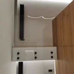 Szklana balustrada schodowa z czarnymi punktowymi mocowaniami, widoczna z dołu, z elementami wystroju wnętrza w tle, takimi jak grzejnik i oświetlenie LED.