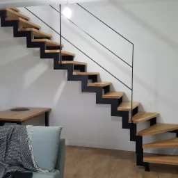 Drewniane stopnie na metalowej konstrukcji w nowoczesnym wnętrzu z minimalistyczną balustradą z czarnych prętów, widoczne z perspektywy salonu z szarą kanapą i drewnianym stołem.