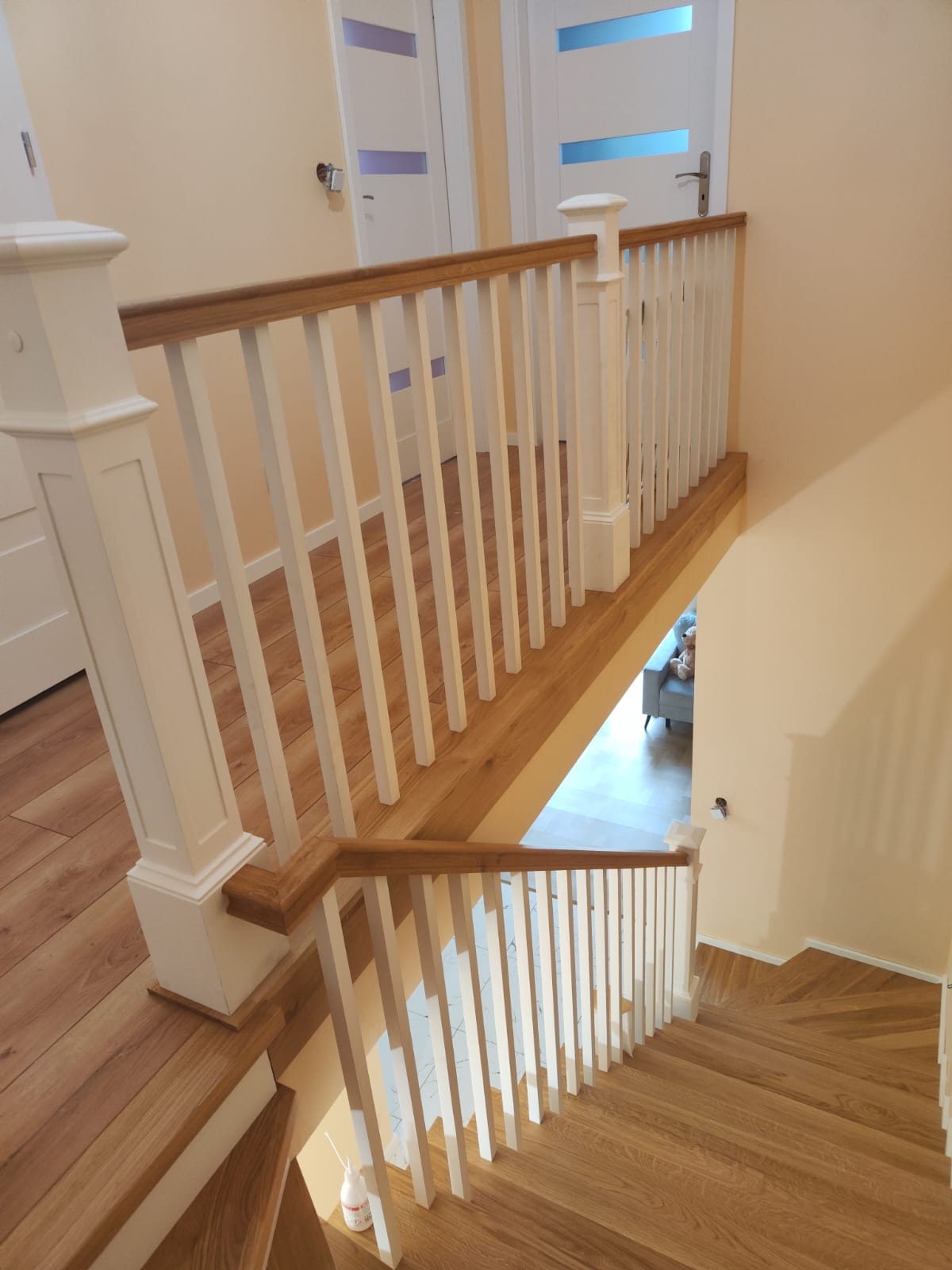 Widok z góry na jasne, dębowe schody z białymi balustradami i poręczą z naturalnego drewna, prowadzące w dół. W tle widoczne drzwi i fragment pokoju z szarą sofą.