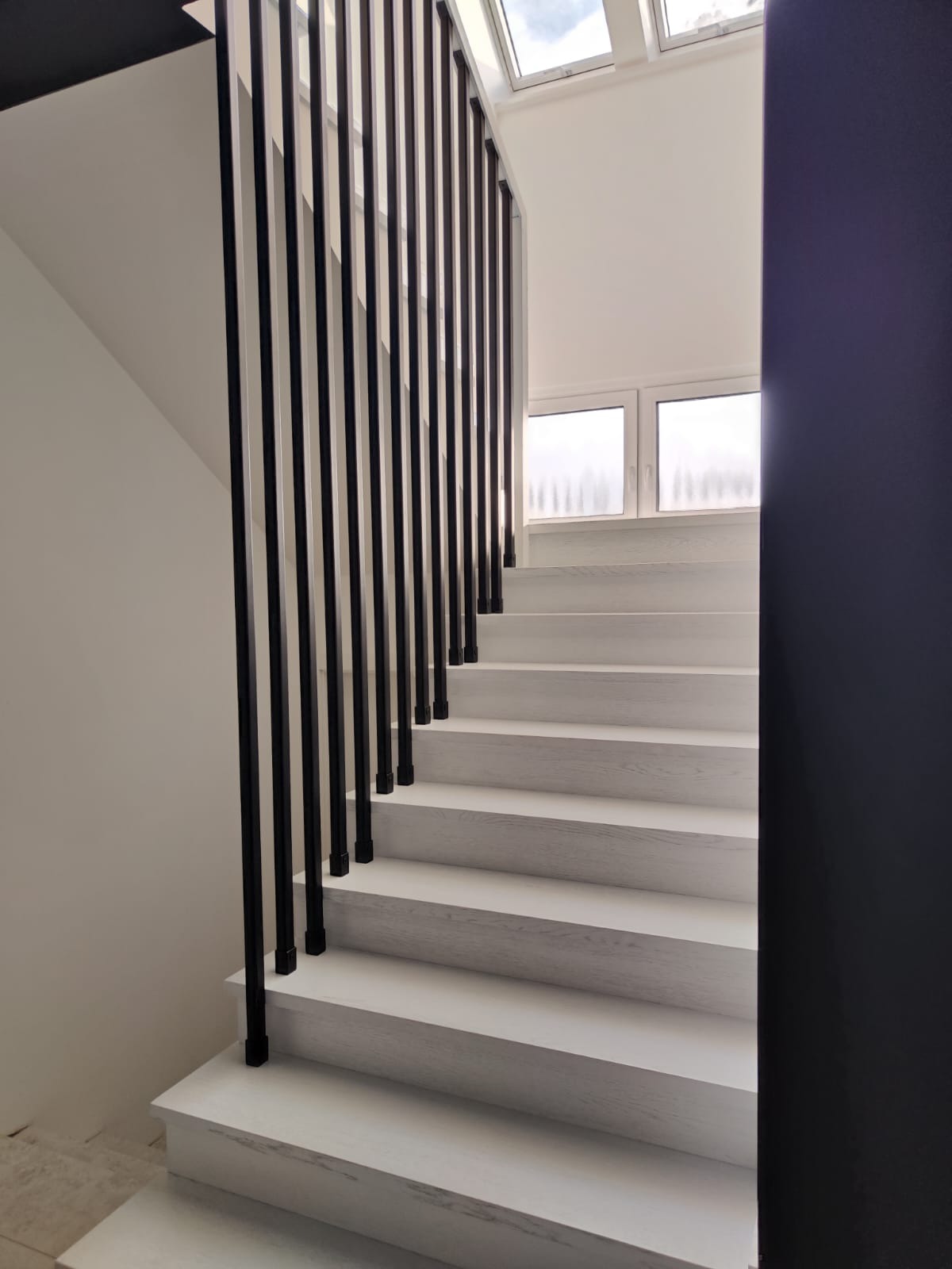 Białe schody z czarnymi, pionowymi balustradami w nowoczesnym wnętrzu z oknem dachowym.