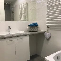 Nowoczesna łazienka z białą umywalką w szafce, grzejnikiem drabinkowym i dekoracyjnym sercem na ścianie z płytek 3D.