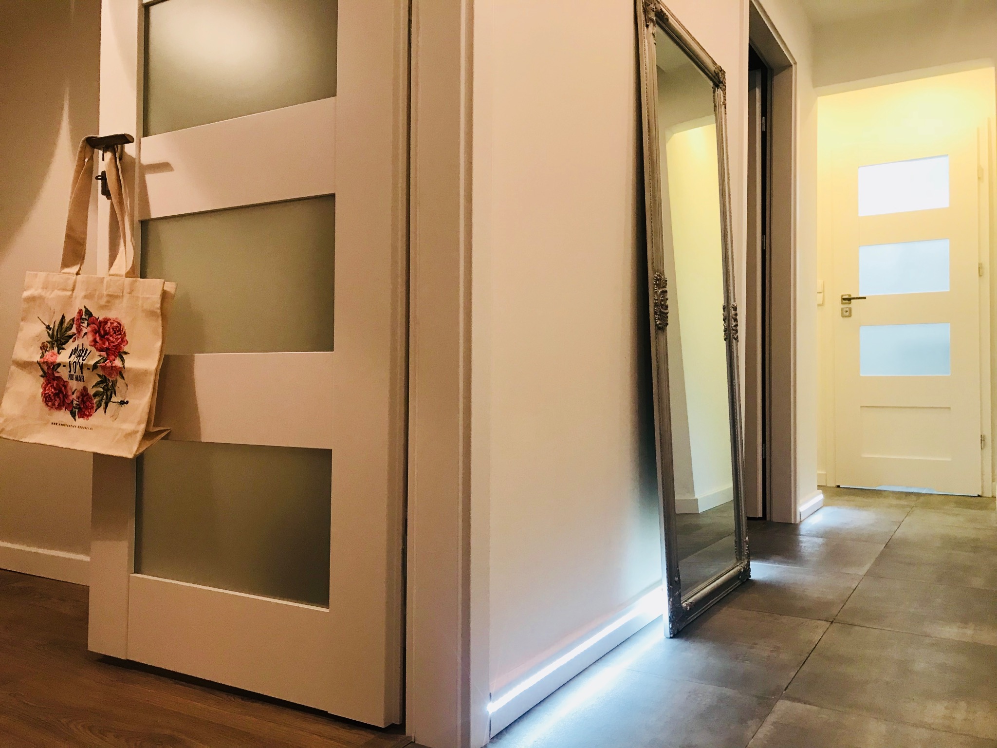 Wnętrze przedpokoju z białymi drzwiami z matowego szkła, torbą z motywem kwiatowym na wieszaku i ozdobnym lustrem opartym o ścianę, podświetlona listwa przypodłogowa.