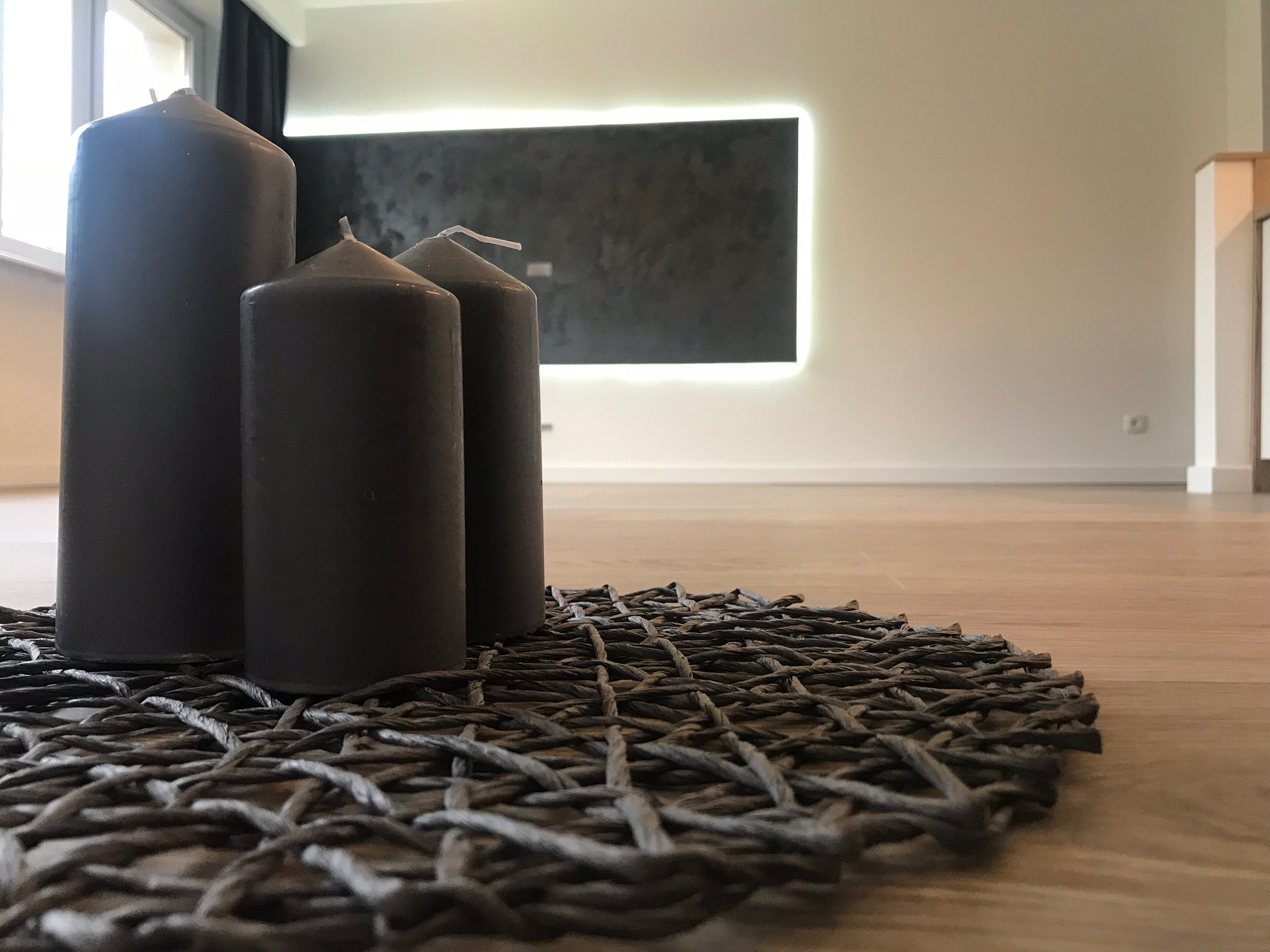 Kompozycja z trzech grafitowych świec na plecionym podkładzie, na tle minimalistycznego wnętrza z drewnianą podłogą i podświetlanym panelem ściennym.