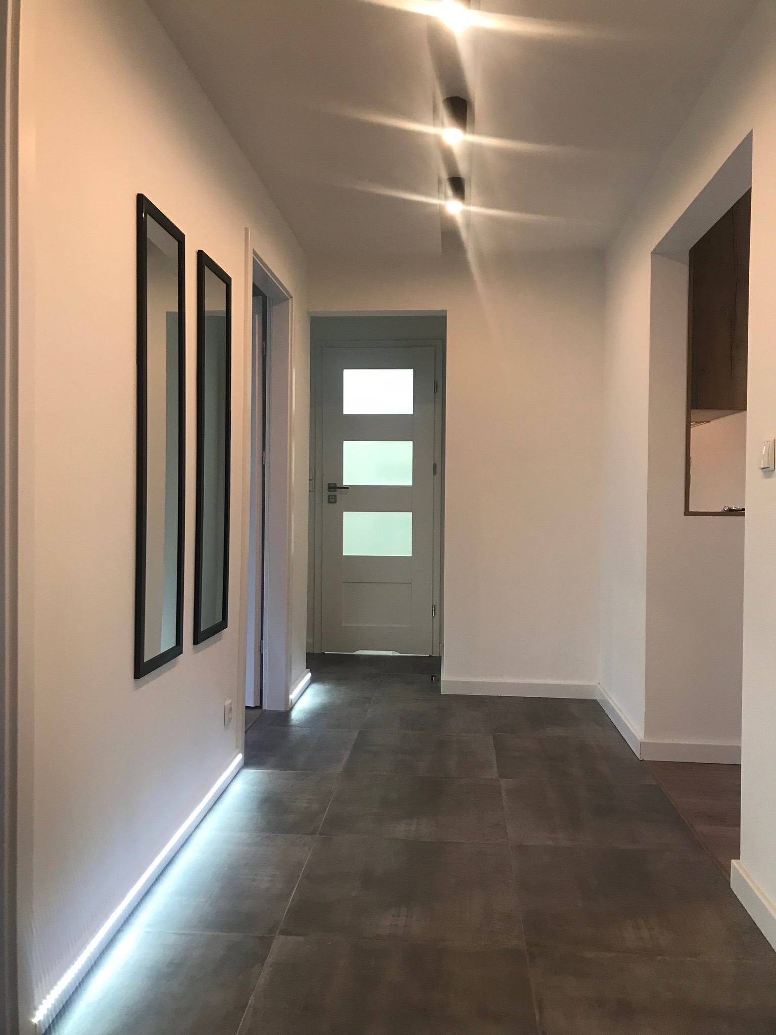 Nowoczesny korytarz z szarymi płytkami podłogowymi, białymi ścianami z podświetleniem LED przy podłodze, dwoma wąskimi lustrami w czarnych ramach oraz trzydrzwiowymi białymi drzwiami na końcu...