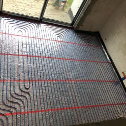 Instalacja ogrzewania podłogowego w nowym budownictwie: niebieskie rury ułożone na czerwonej siatce, widok z góry na surowy stan wykończenia.