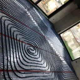 Rozłożona instalacja ogrzewania podłogowego z czarnych rurek na srebrnej macie z czerwoną siatką, widoczne okna i surowy stan wykończenia pomieszczenia.