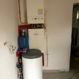 Nowo zainstalowany, wiszący, biały piec gazowy z podłączonym zasobnikiem ciepłej wody, rurami miedzianymi i czerwonymi rurami instalacji CO, w surowym pomieszczeniu z odsłoniętymi przewodami...