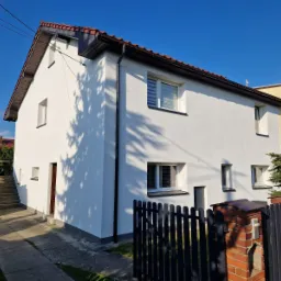 Biały dom jednorodzinny po remoncie elewacji, widoczny z zewnątrz, z czarnymi rynnami i brązową stolarką okienną oraz drewnianym dachem z czerwoną dachówką, częściowo zacieniony przez drzewa.