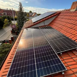 Panele fotowoltaiczne zamontowane na dachu domu z czerwonej dachówki, widoczne z perspektywy dachu; promienie słoneczne odbijają się od powierzchni paneli.