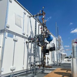 NOWY KLIMAT SPÓŁKA Z OGRANICZONĄ ODPOWIEDZIALNOŚCIĄ - Rozbudowana instalacja rur i zaworów na dachu budynku z białymi panelami, błękitne niebo w tle, widoczne elementy konstrukcyjne dachu i zabezpieczenia instalacji.