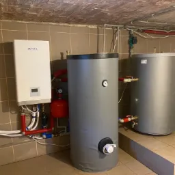 Biały wiszący piec grzewczy marki Seyra z podłączonymi rurami, obok dwa szare cylindryczne zbiorniki na wodę w kotłowni wyłożonej brązowymi płytkami.