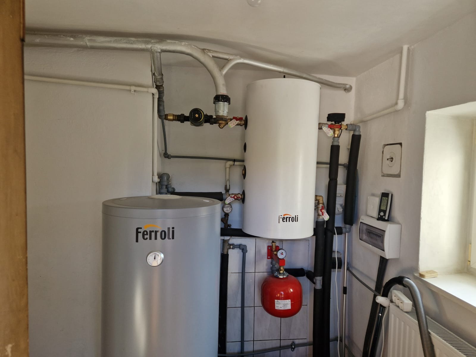 Dwa zbiorniki buforowe Ferroli w kotłowni, instalacja z rurami, zaworami i manometrami, czerwony zbiornik przeponowy, skrzynka elektryczna, grzejnik żeberkowy przy oknie.