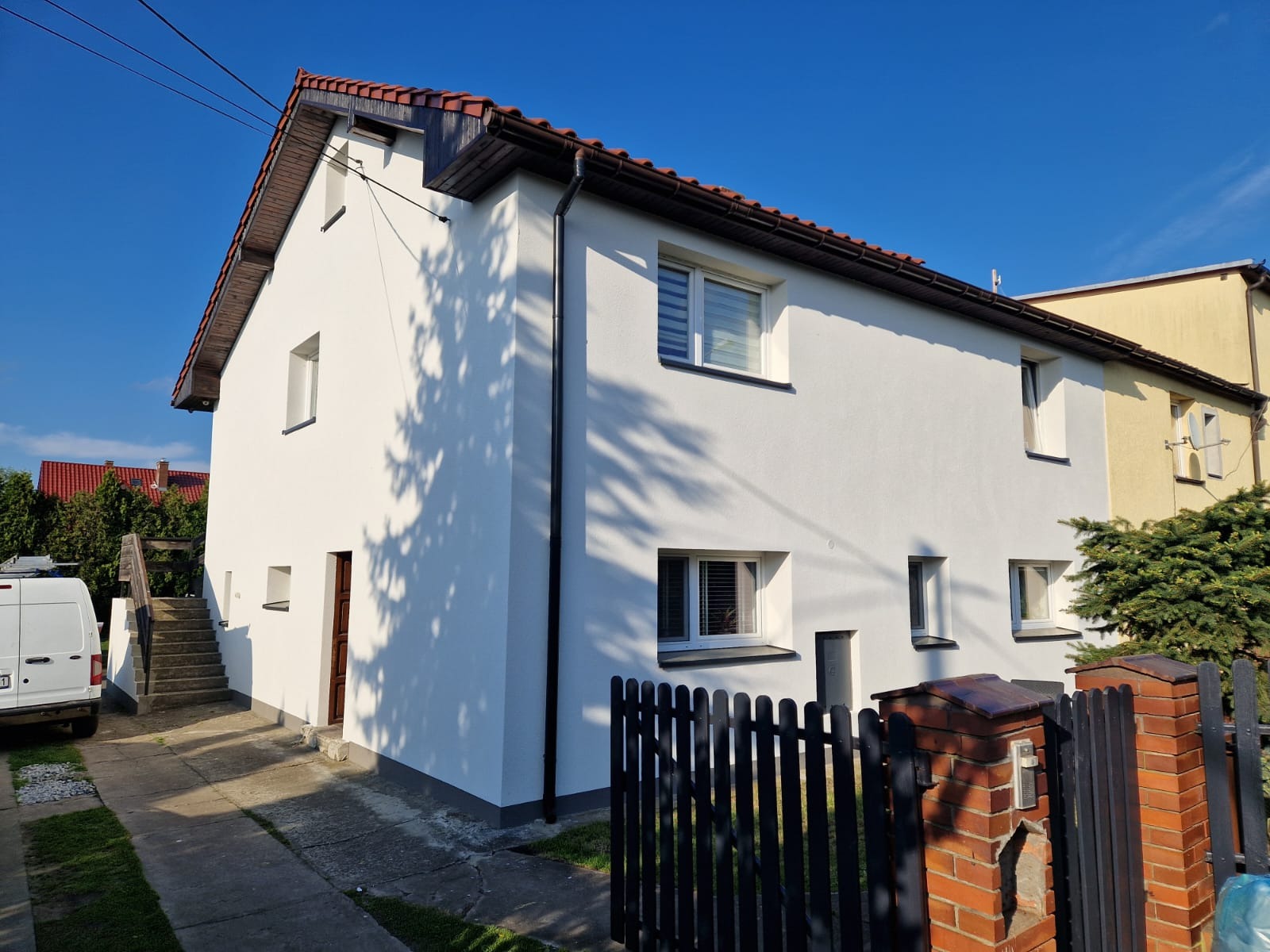 Biały dom jednorodzinny po remoncie elewacji, widoczny z zewnątrz, z czarnymi rynnami i brązową stolarką okienną oraz drewnianym dachem z czerwoną dachówką, częściowo zacieniony przez drzewa.