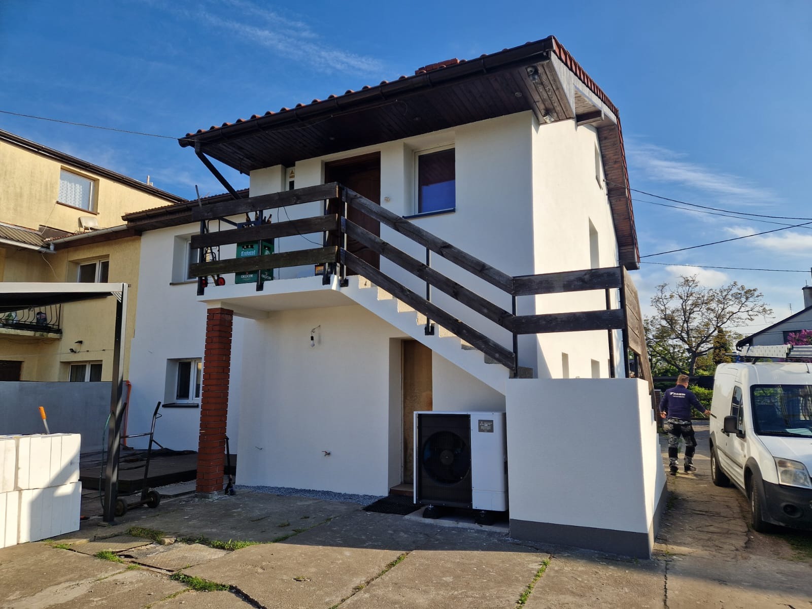 Biały, dwukondygnacyjny dom z zewnętrzną pompą ciepła Mitsubishi ustawioną przy ścianie, obok schodów wejściowych i zaparkowanego białego samochodu dostawczego, na tle błękitnego nieba.