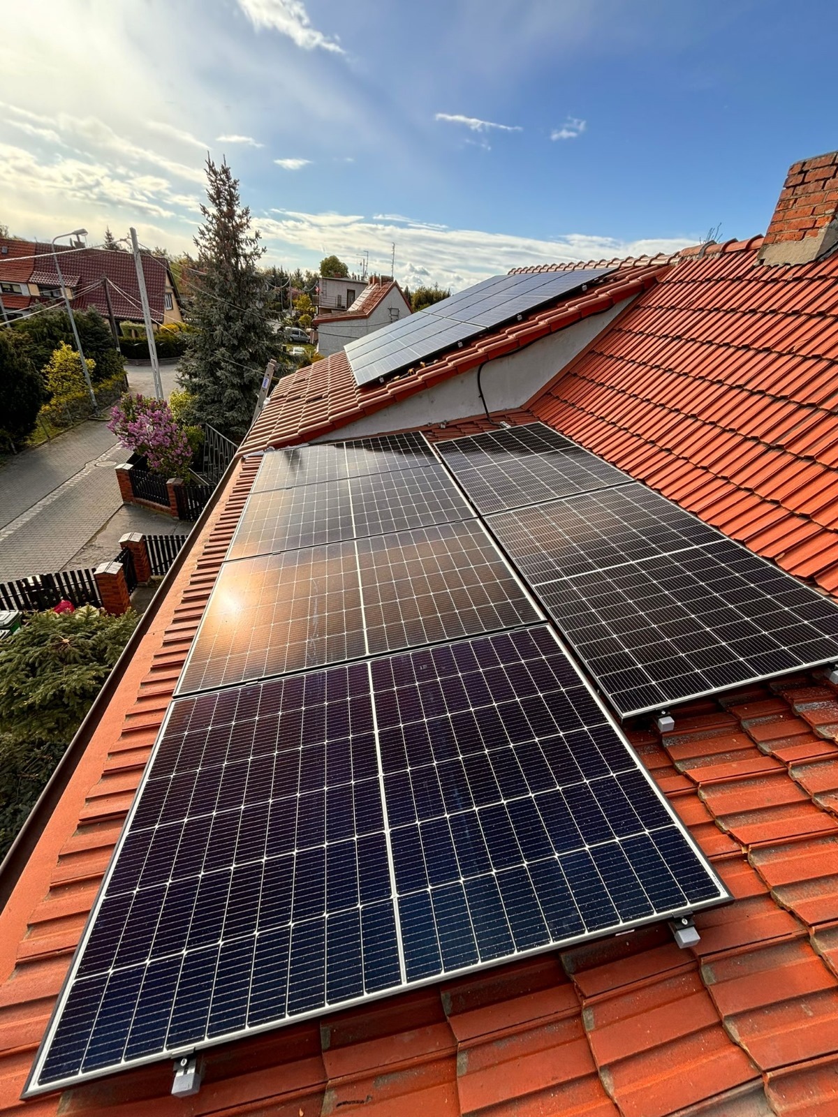 Panele fotowoltaiczne zamontowane na dachu domu z czerwonej dachówki, widoczne z perspektywy dachu; promienie słoneczne odbijają się od powierzchni paneli.