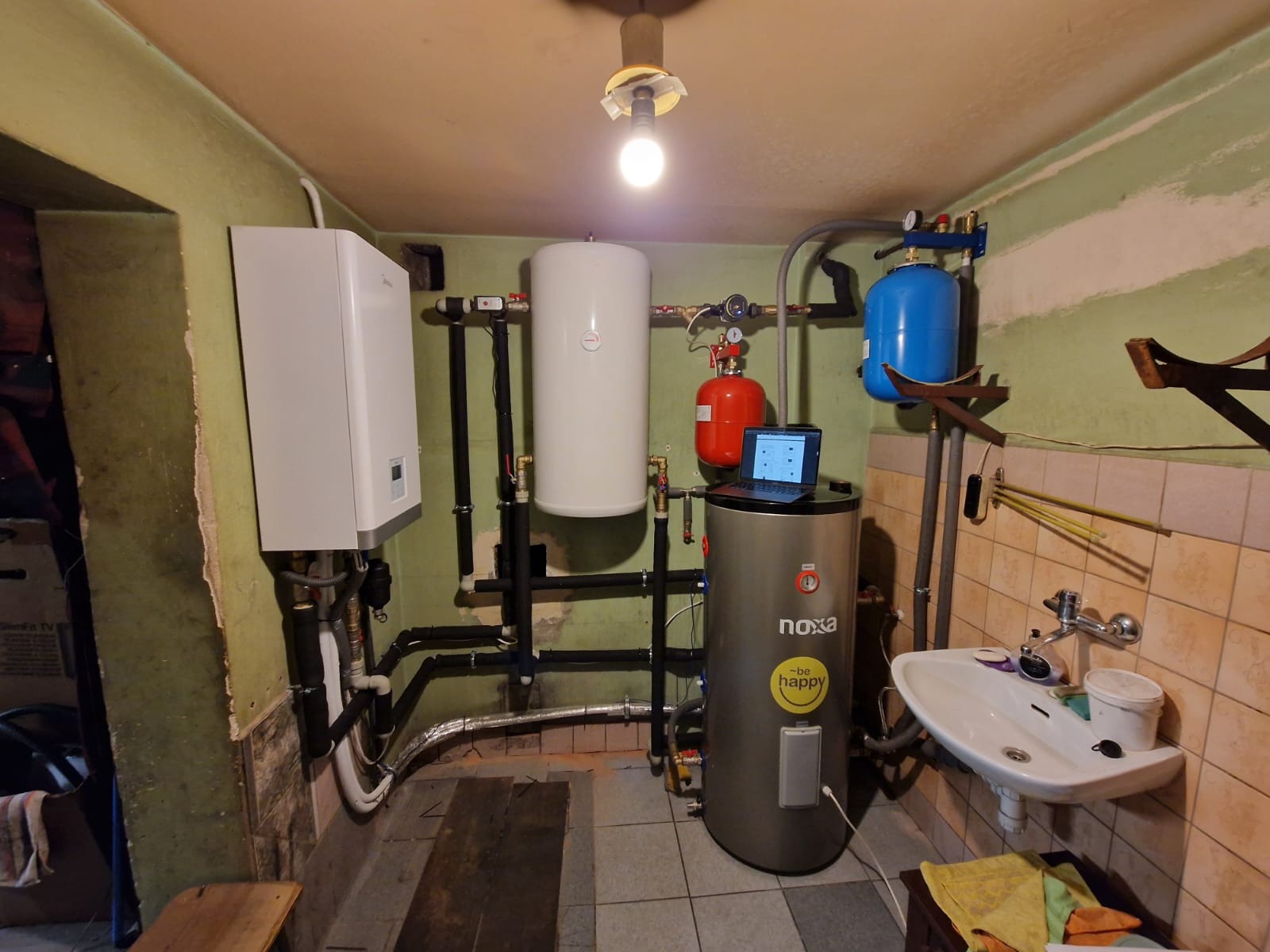 Kotłownia w piwnicy z zainstalowanym piecem, zbiornikiem buforowym, naczyniami przeponowymi i laptopem na zbiorniku, widoczne rury instalacyjne i umywalka.