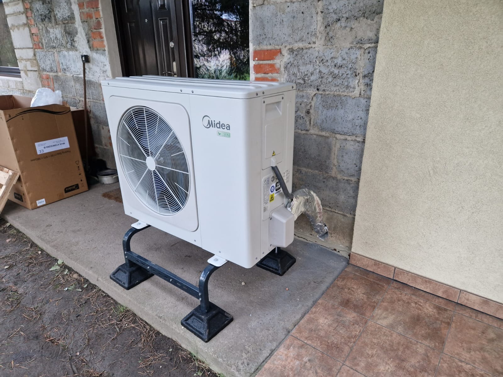 Zewnętrzna jednostka klimatyzacji Midea na czarnym, metalowym stojaku, ustawiona przy szarej ścianie z cegieł i beżowej elewacji, widoczna kartonowa paczka w tle.
