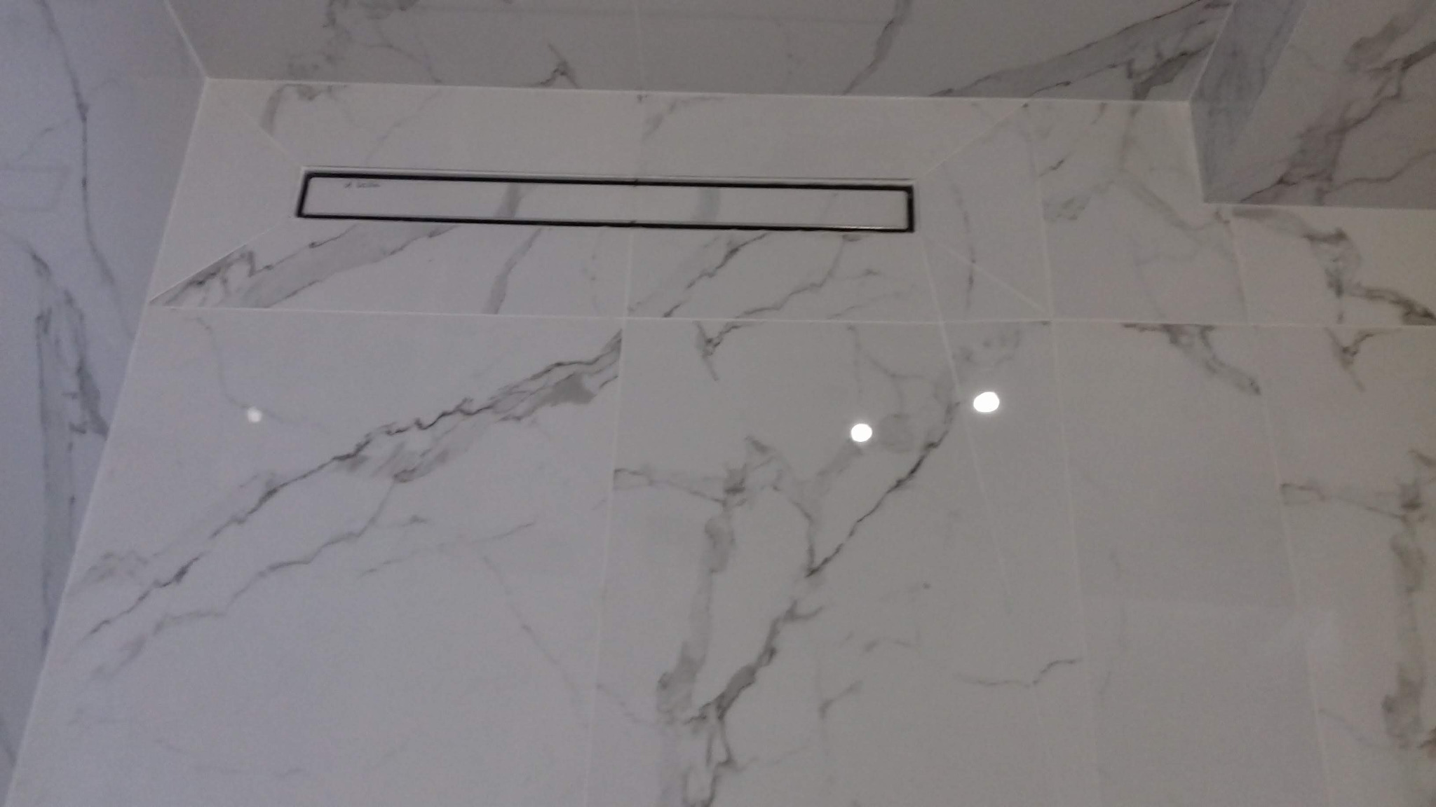 Ściana wykończona białymi płytkami imitującymi marmur z wbudowanym odpływem liniowym w kabinie prysznicowej.