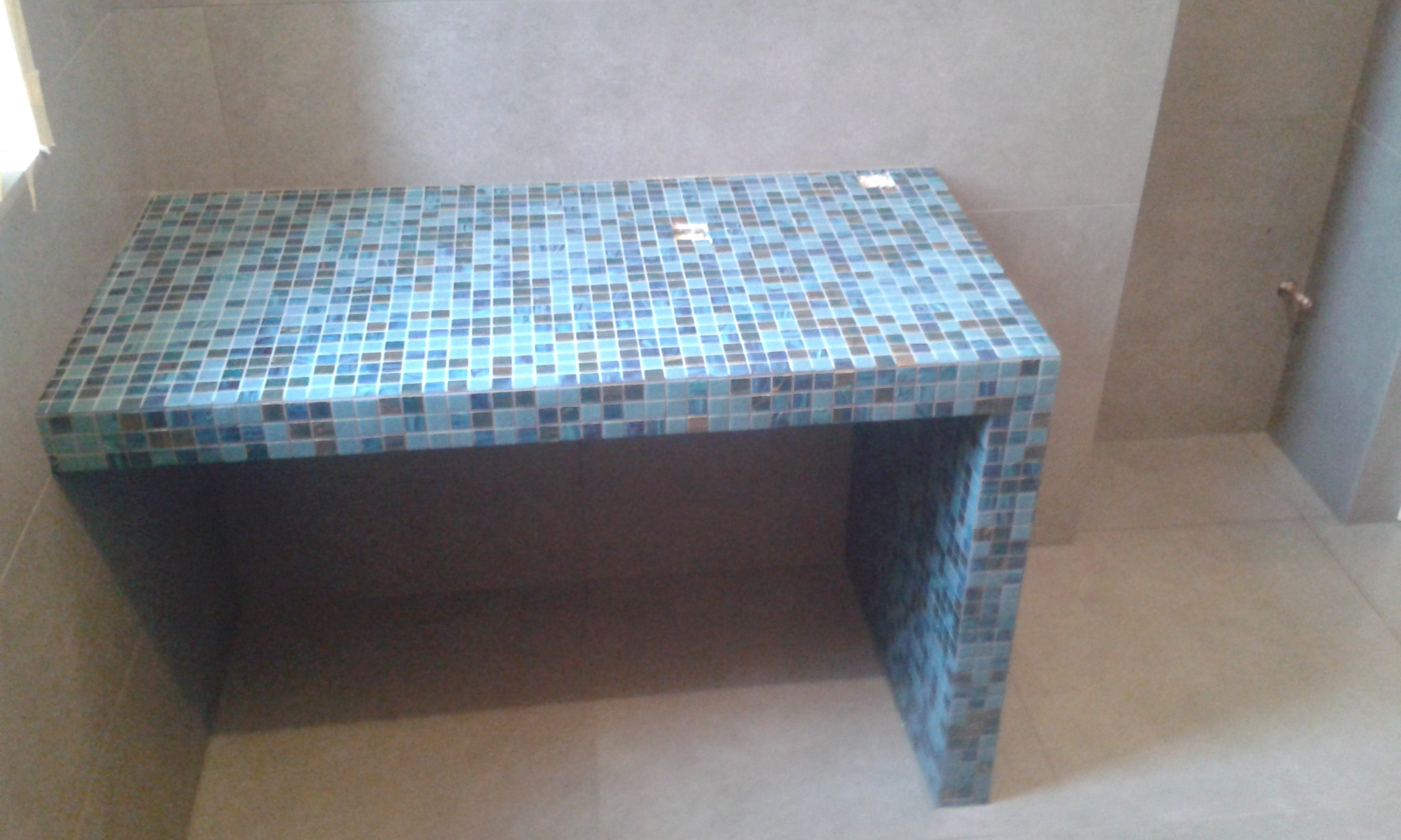 Narożny blat łazienkowy wykończony mozaiką w odcieniach błękitu i brązu, dopasowany kolorystycznie do płytek podłogowych i ściennych.