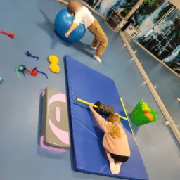Dwoje dzieci ćwiczy w sali rehabilitacyjnej: jedno na dużej niebieskiej piłce, drugie na niebieskim materacu z wałkiem i kijkami.
